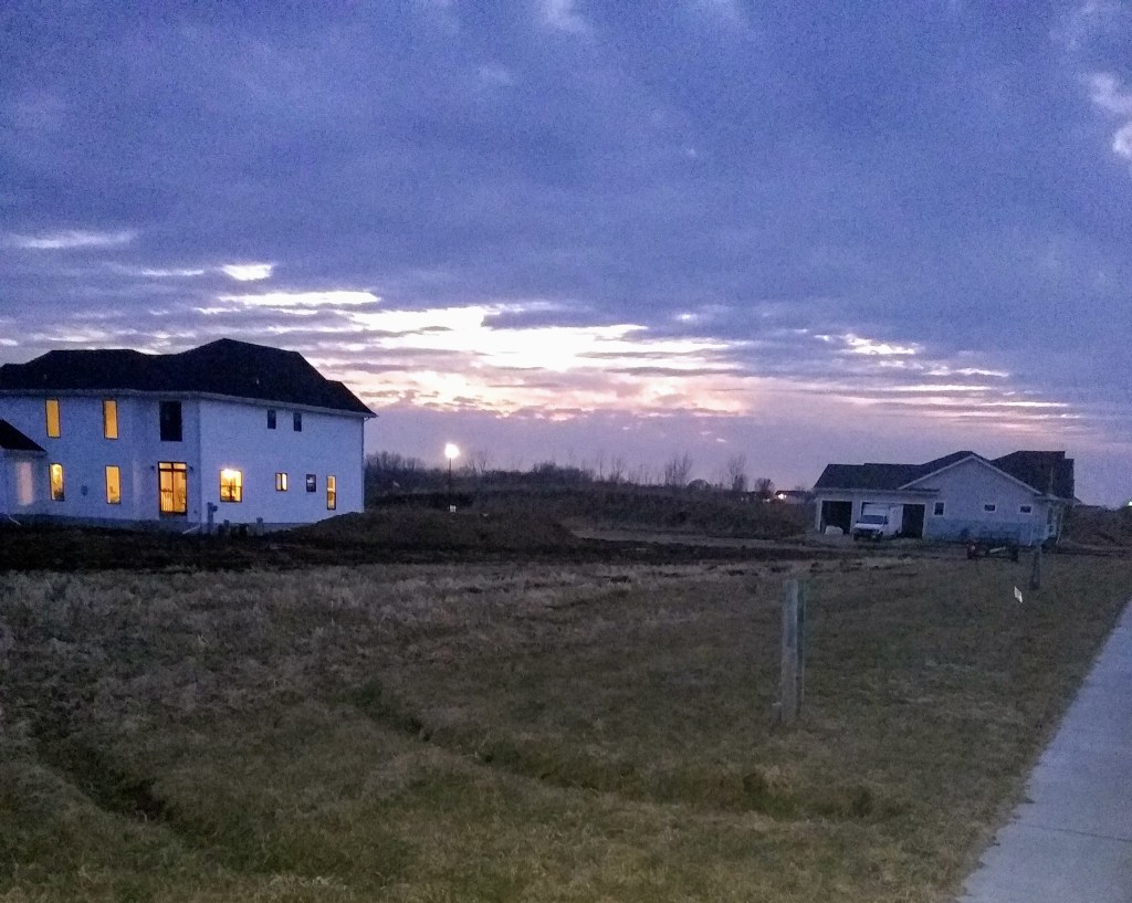 Recently built houses, one with windows brightly lit, the other dark with a truck parked in the driveway. The sun is setting behind a bank of purple clouds in the background between the two houses.